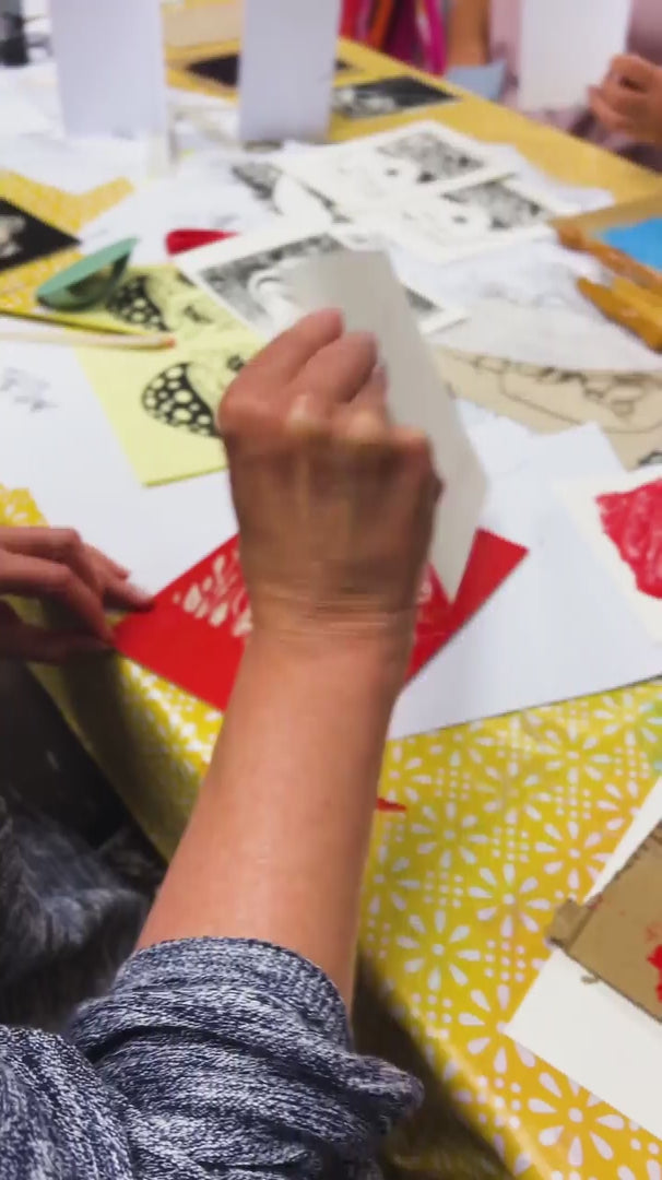 Beginner lino printing workshop at Maker Studio Eastbourne,   tutor demonstrating how to carve a linocut block on an ink-ready workbench, surrounded by printmaking tools, brayers, carving tools and ink rollers, hands-on creative craft class for adults learning relief printing techniques in a friendly community studio setting