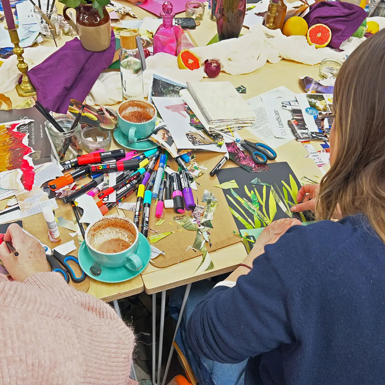 Two people working on art projects at a table with coffee cups, markers, and scissors.